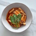 Delicious bowl of spicy Korean tteokbokki and noodles topped with fresh greens and sesame seeds.
