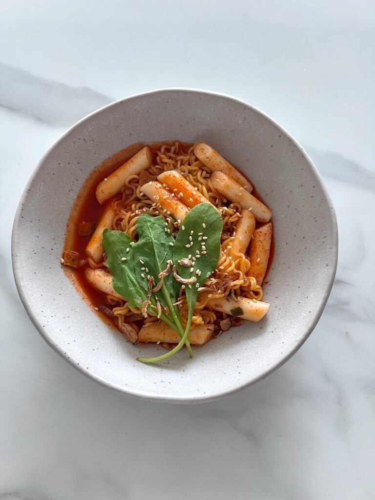 Delicious bowl of spicy Korean tteokbokki and noodles topped with fresh greens and sesame seeds.